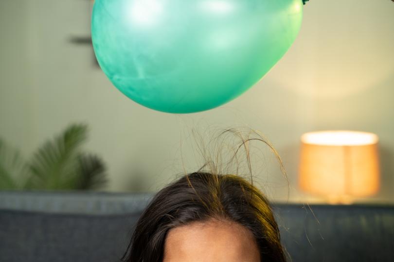 an unseen hand holds a green latex balloon over the top of a child's head, and her hair is reaching up to the balloon through static electricity