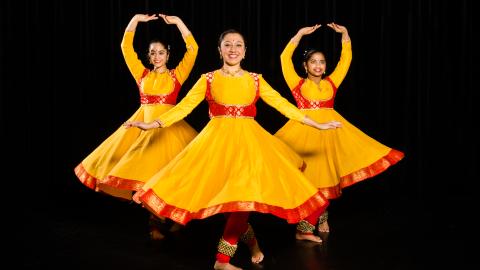 Three female dancers twirl wearing bright yellow and red costumes from India