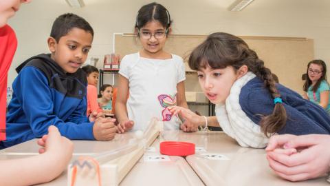 A small group of children watches a marble roll down a wooden track