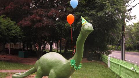 a large dinosaur statue has balloons tied to it and wears a party hat
