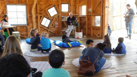 a group of kids and adults are sitting on the floor of the Discovery Treehouse listening to a man seated in the corner read a book