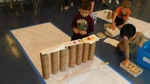 a boy kneels on the floor with a line of cardboard tubes with cardboard and papers across the top