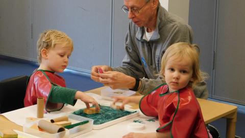 two small kids sit at a table with an older man. Each has a piece of paper in front of them and the kids are dipping stamps into paint