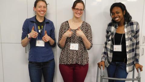 Three adult women wearing nametags stand together; two give two "thumbs up," one is standing holding a walker. All three are smiling!