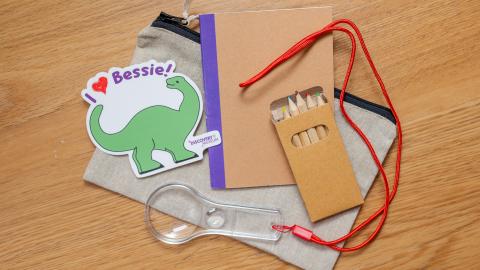a small notebook, magnifying glass, pencil and dino sticker on top of a small canvas bag