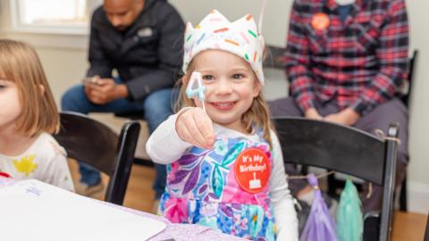 a young girl wearing a birthday crown holds up a candle in the shape of a number 4