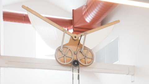 a young child turns a crank attached to a set of "wings" above, as her father looks on