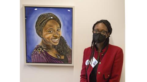 Scientist Ellice Patterson wears glasses, a black face mask, and a red jacket and poses next to a painting of herself in the Discovery Museum