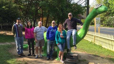 6 adults stand by or sit on a large dinosaur statue