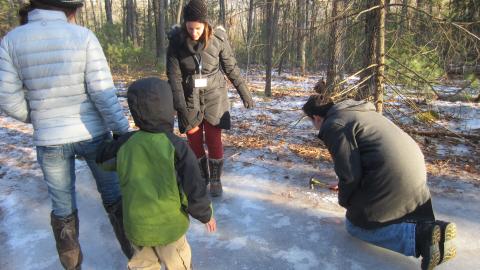 Three adults and a child are wearing coats in the woods looking at a patch of ice on the ground