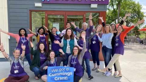 A group of Discovery Museum employees pose in front of the Museum door with hands in the air and holding a poster that says "We got a Cummings Grant" 