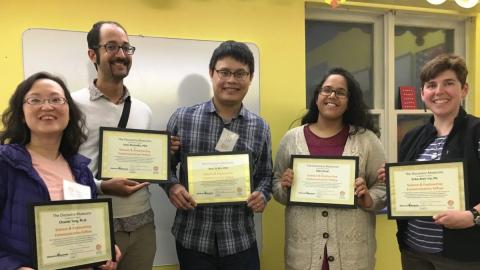 Five adults stand together, smiling, each holding a framed certificate