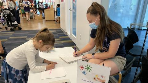 a child stands in front of a table that a woman is seated behind, and draws a picture