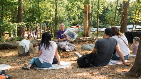 Adults and kids sit on the ground in the woods listening and watching a man read a picture book.