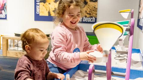 two children stand at a table full of sand, pouring sand through funnels and into dishes