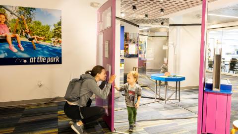 a mom peers through a window in a mirrored exhibit to her child on the other side