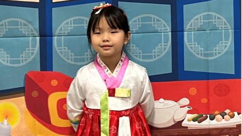 A girl wearing traditional clothes stands in front of a backdrop made to look like the inside of a Korean home