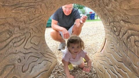 a young child crawls through a playstructure tunnel at a playground, while her grandfather looks on