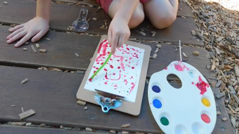 A child sits on the ground holding a paintbrush to a paper on the ground and a paint palette of different colors is on the ground next to it. Only the child's hands are visible