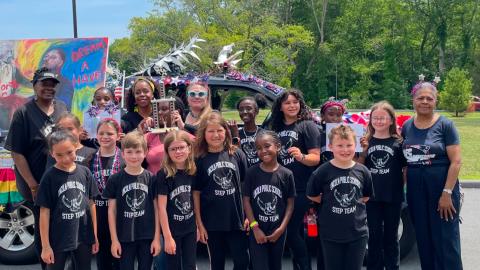 A group of 12 to 15 kids wearing black tshirts and pants with a couple of adults pose outside on a sunny day