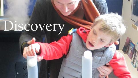 a boy's mother holds him up out of his wheelchair as he explores putting ping pong balls over air tubes