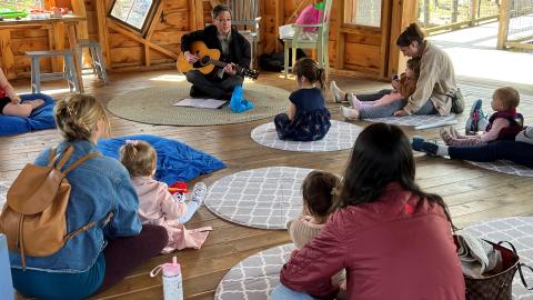 A man sits on the floor playing guitar and there are women and children sitting on the floor around him.