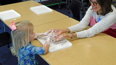 A woman sits across the table from a little girl with blond hair and a blue dress. They have their hands in a tray of shaving cream that is between them. 