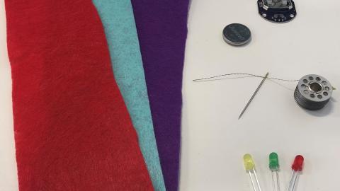 Three pieces of felt in red, blue, purple. A needle and thread. Button battery and LED light pieces in yellow, green, and red
