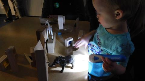 A child shines a flashlight on toy figures and blocks on a table in a dark room
