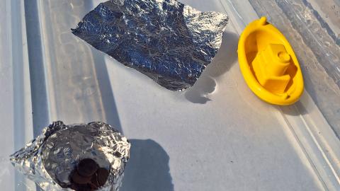 a clear plastic container full of water on a table. Floating on the water are two pieces of foil, one has pennies on it, and a little toy boat
