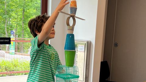 A boy is stacking various items (cups, cardboard, baskets) into a tower.