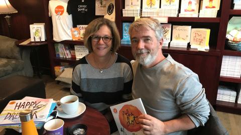 a man and a woman sit in a coffee shop; the man holds up a book that he wrote
