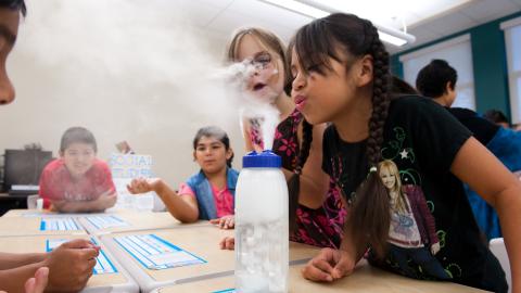 a girl blows on white vapor coming out of a bottle in an experiment