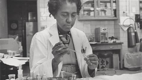 A black and white photo of a black woman in a lab coat sitting in a lab holding a test tube.