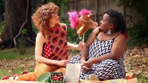 A white woman and black woman are sitting on a blanket outside smiling at a colorful puppet of a chicken between them.