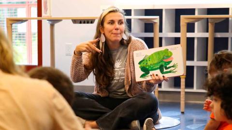 a woman sits on the floor reading a picture book to children, using sign language