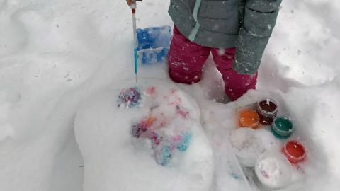 A child shown from the waist down dressed in snow gear and standing in snow up to their knees is dripping paint on to the snow.