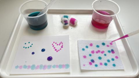 a white tray is on a white table holding a plastic cup of blue water and a plastic cup of red water. There are two napkins with dots of blue and red and a plastic pipette with red water