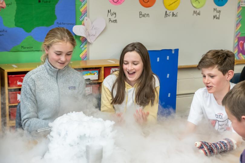 three middle school-aged students at a table are wowed by condensation billowing from a container where dry ice has been dropped into water