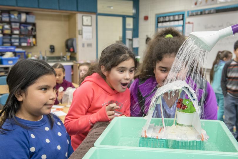 three students watch as an adult pours water from a watering can onto the animal shelter they created to keep a toy animal dry. One girl has her fingers crossed as the water hits the shelter.