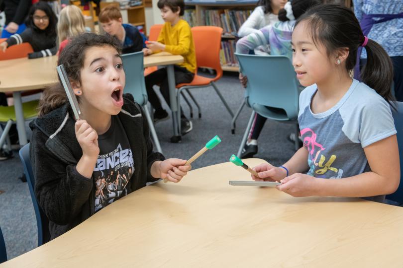 two students sit at a desk, one holds a vibrating tuning fork near her ear and is yelping with surprise!