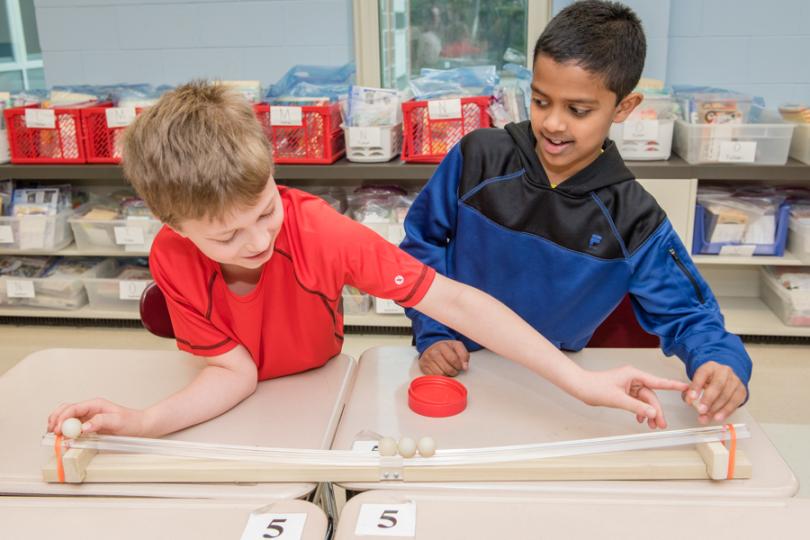 two boys in a classroom roll small steel balls down a tabletop wooden ramp