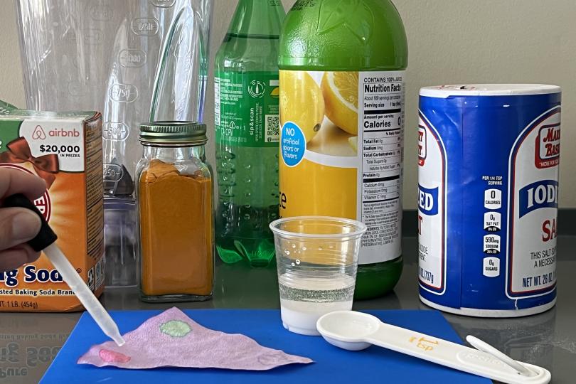 a hand uses a dropper to put drops of liquid onto pink paper on a table; behind are common kitchen items such as salt, lemon juice, turmeric, and baking sods