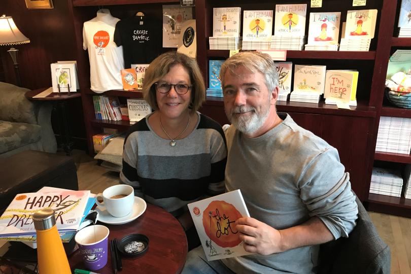 a man and a woman sit in a coffee shop; the man holds up a book that he wrote