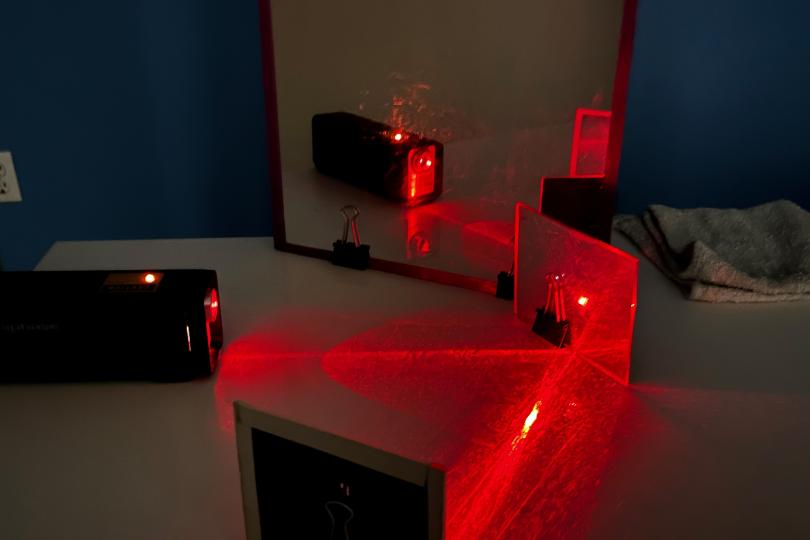 a dark photo showing red laser light reflected off a mirror and other reflective objects on a table