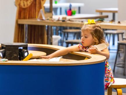 a young girl reaches onto a table that has spinning plates with sand on them; she makes a pattern