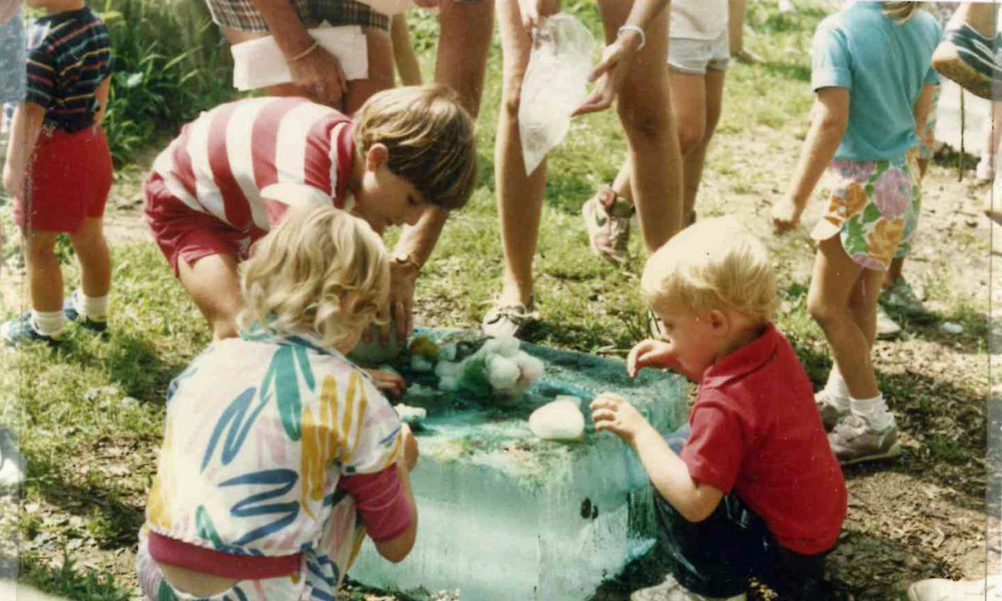 several children stoop to explore a very large, partially melted, 300lb block of ice that is sitting on the ground