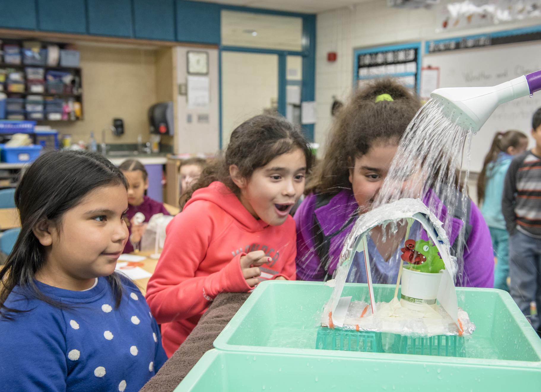three students watch as an adult pours water from a watering can onto the animal shelter they created to keep a toy animal dry. One girl has her fingers crossed as the water hits the shelter.