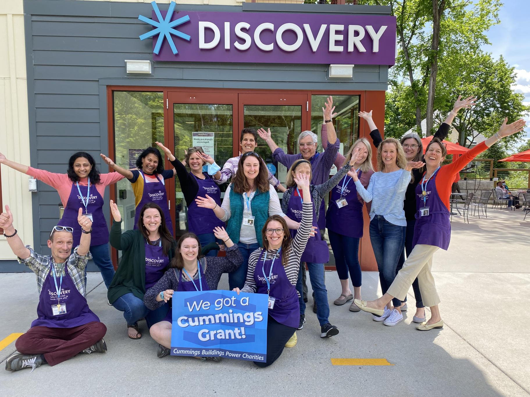 A group of Discovery Museum employees pose in front of the Museum door with hands in the air and holding a poster that says "We got a Cummings Grant" 