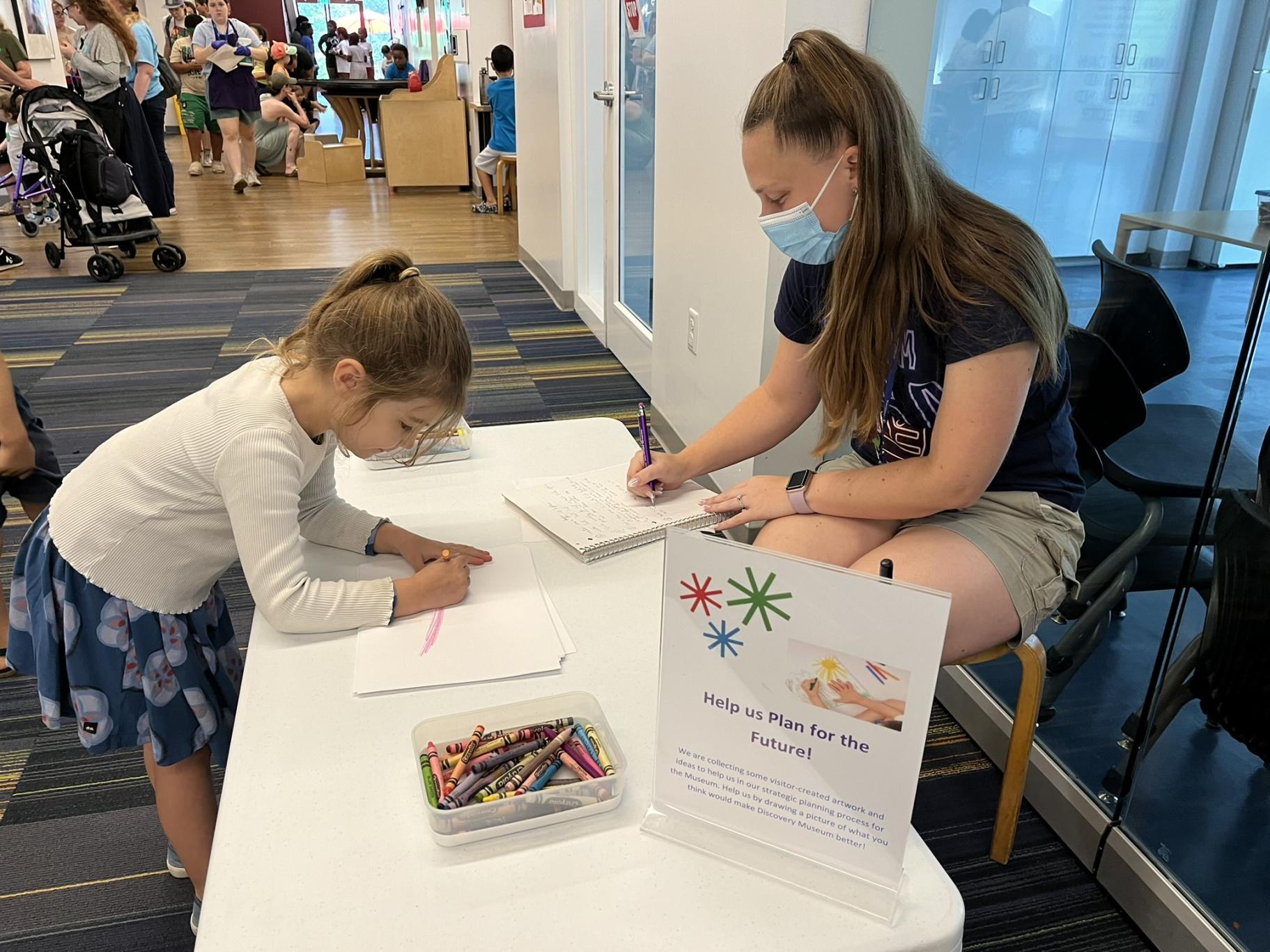 a child stands in front of a table that a woman is seated behind, and draws a picture